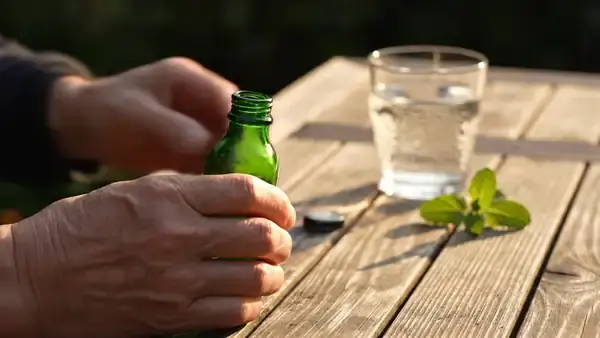 A man's hands tipping two dark liquid capsules from a small green bottle beside a glass of water and a sprig of spearmint